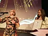 To kvinner på scene, en taler i mikrofon, den andre holder et innrammet diplom.