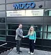 To personer håndhilser foran MDCO-skiltet på en mørk glassfasade.