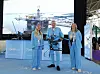 To kvinner og en mann står på en utstilling. Mannen holder en stor drone. I bakgrunnen en storskjerm som viser en oljeplattform liggende til kai i en havn.