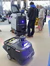 En profesjonell liten feierobot vises på stand under en renholdsmesse. I bakgrunnen ses flere mennesker som ser på en annen rengjøringsmaskin.