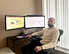En mann sitter ved siden av et skrivebord. Oppå skrivebordet står to dataskjermer som viser et bykart.