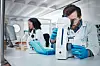 Laboratoriearbeider med blå hansker bruker mikroskop i et lyst laboratorium.